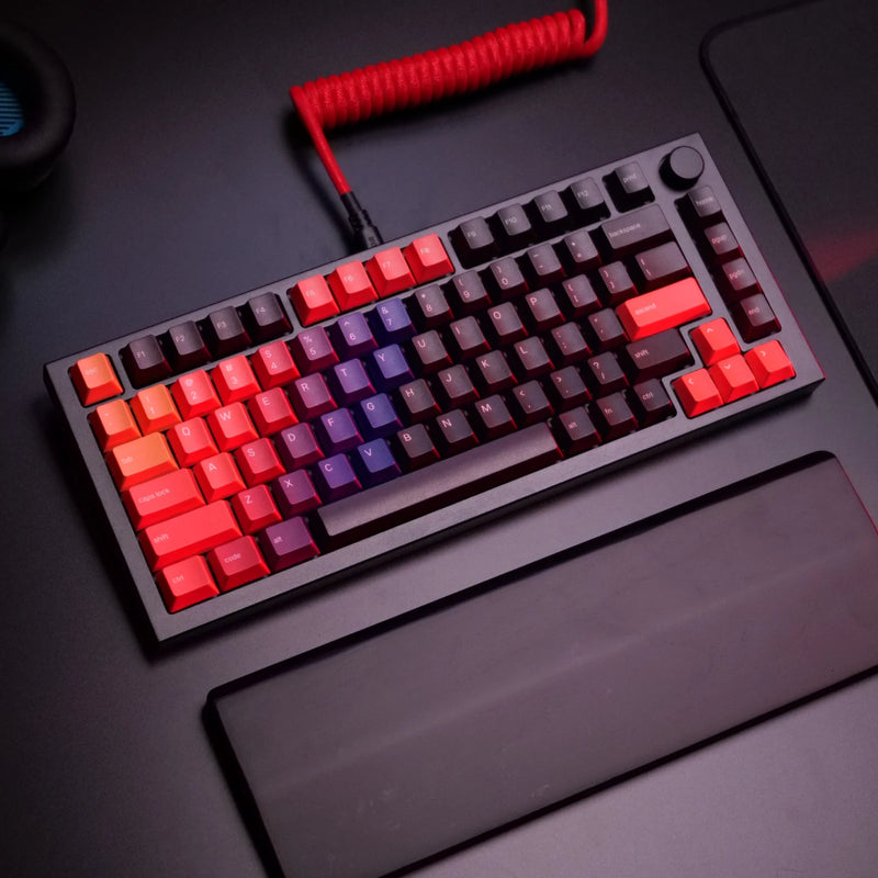 A GMMK PRO in Black Slate with GPBT Celestial Fire keycaps and a Crimson Red coiled cable connected is shown on a dark desk. A black wooden wrist rest is positioned in front of the keyboard.