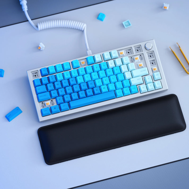 A GMMK PRO Barebones in White Ice with GPBT Ocean keycaps is shown on a desk with some keycaps removed, exposing the switches underneath. A black wrist rest is positioned in front of the keyboard, and a Ghost White coiled cable is connected to it. Additional keycaps and tools are scattered around the desk.