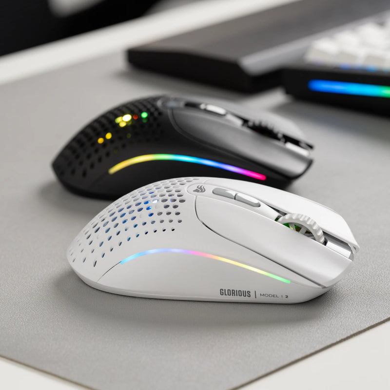 A white Glorious Model I 2 Wireless gaming mouse with RGB lighting is placed in front of a black Model I 2 Wireless on a mousepad.