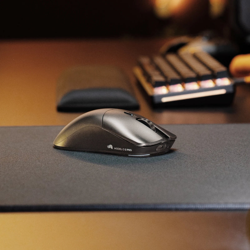 A side view of a Model O 2 PRO Series Wireless Mouse on a dark mouse pad, with a black GMMK 2 65% and other desk accessories blurred in the background.
